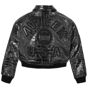May include: Black leather jacket with a crocodile skin texture. The back of the jacket features the brand name "AVIREX USA" in raised lettering and a circular emblem. The jacket has a ribbed collar and cuffs.