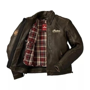 Puede incluir: Chaqueta de cuero marrón con forro a cuadros rojos y negros en el interior. La chaqueta tiene cuello, cremallera y la palabra "Indian" en letras blancas. Se ve un parche en la manga izquierda.