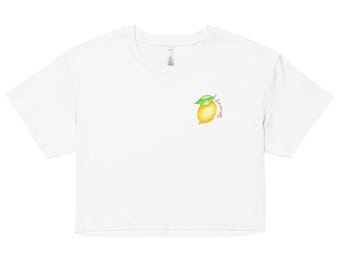 Op de afbeelding: Witte crop top met een aquarel citroen ontwerp, een groen blad en het woord "Lemonade" in cursief. De top heeft een ronde hals en korte mouwen. Een casual, zomerse stijl.