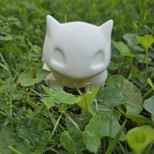Puede incluir: Una figura de cerámica blanca de una criatura felina con grandes orejas y una carita sonriente. La figura está colocada en un lecho de hierba y hojas verdes, creando un entorno natural. El personaje es popular.