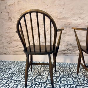 May include: A dark brown wooden chair with a curved back and vertical spindles. The chair has armrests and a seat. The chair is set against a textured white wall and sits on a patterned tile floor.