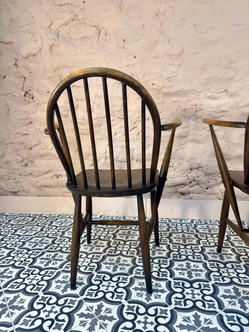 May include: A dark brown wooden chair with a curved back and vertical spindles. The chair has armrests and a seat. The chair is set against a textured white wall and sits on a patterned tile floor.