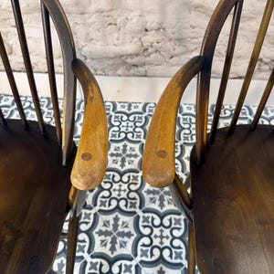 May include: Two wooden chairs with curved backs and armrests. The chairs have a dark brown finish with lighter wood armrests. The chairs are positioned on a patterned tile floor with a neutral-colored wall in the background.