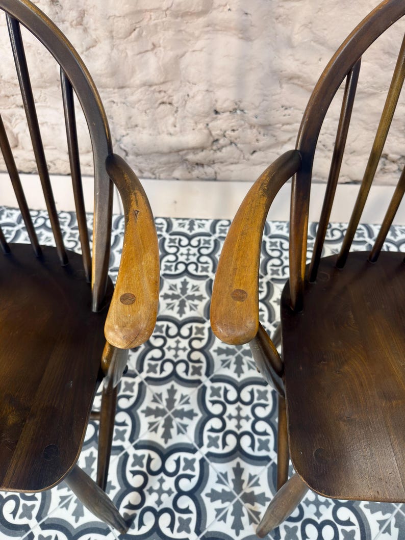 May include: Two wooden chairs with curved backs and armrests. The chairs have a dark brown finish with lighter wood armrests. The chairs are positioned on a patterned tile floor with a neutral-colored wall in the background.