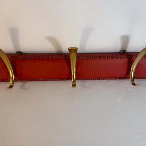 May include: A red wall-mounted coat rack with three brass hooks. The rectangular wooden base is covered in red material and adorned with small, evenly spaced rivets. The curved hooks provide a stylish storage solution.