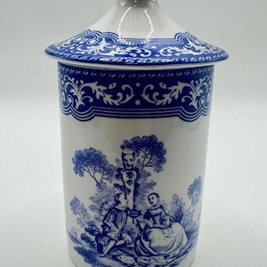 Puede incluir: Un tarro de cerámica blanco con un diseño toile de Jouy azul y blanco. El tarro tiene una tapa con un borde azul decorativo y un pomo. El cuerpo del tarro presenta una escena pastoral con figuras y árboles.