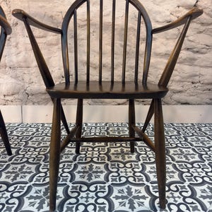 May include: Dark brown wooden chair with a curved back and armrests. The chair has a spindle back design and a sturdy frame. The chair is set against a textured, light-colored wall and sits on a patterned tile floor.