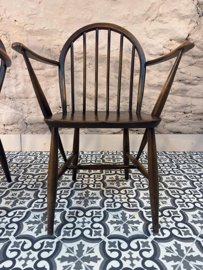 May include: Dark brown wooden chair with a curved back and armrests. The chair has a spindle back design and a sturdy frame. The chair is set against a textured, light-colored wall and sits on a patterned tile floor.