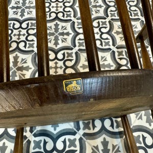 May include: Close-up of a dark brown wooden chair with a spindle back. The chair has a rectangular seat and a label that reads "THIS IS ERCOL PRODUCTIONS". The chair is set against a patterned tile background.