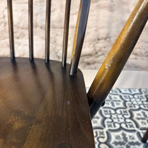 May include: Close-up of a dark brown wooden chair with a spindle back. The chair has a curved backrest and a seat with a circular detail. The wood grain is visible, and the chair appears to be well-worn.