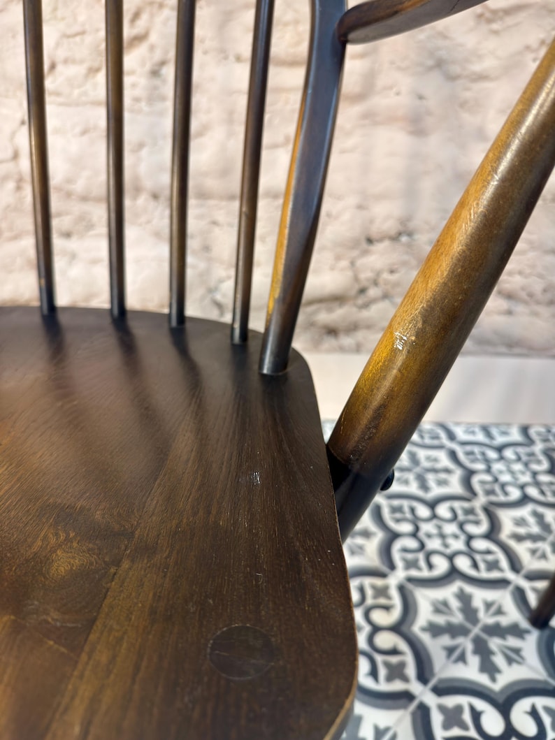 May include: Close-up of a dark brown wooden chair with a spindle back. The chair has a curved backrest and a seat with a circular detail. The wood grain is visible, and the chair appears to be well-worn.