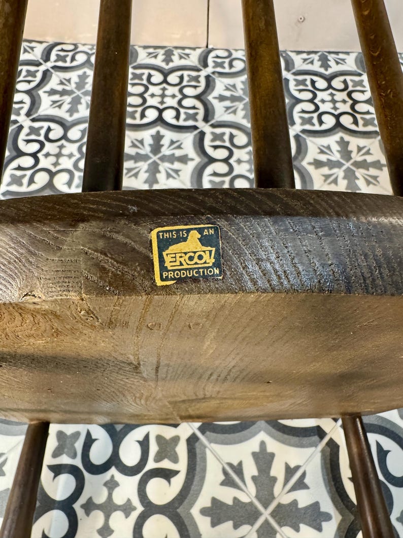 May include: Close-up of a wooden chair seat with a dark finish. The seat has a visible wood grain texture and is supported by multiple curved legs. A sticker on the seat reads "THIS IS AN ERCOL PRODUCTION".