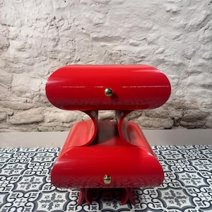 Könnte beinhalten: Ein auffälliger roter Nachttisch oder Beistelltisch im Mid-Century-Modern-Stil. Er hat zwei abgerundete, horizontale Abschnitte mit messingfarbenen Knöpfen. Der Hintergrund ist eine weiße Backsteinmauer.