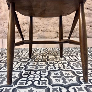 May include: A dark brown wooden chair with a curved seat and spindle back. The chair has four legs and a horizontal support bar. The chair is set against a textured white brick wall and sits on patterned floor tiles.