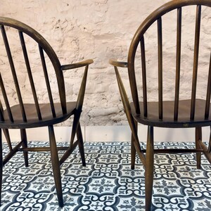 May include: Two dark wooden chairs with curved backs and armrests. The chairs have vertical back supports and are set against a textured white wall. The chairs are on a patterned tile floor with shades of gray and blue.