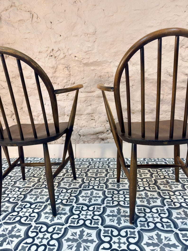 May include: Two dark wooden chairs with curved backs and armrests. The chairs have vertical back supports and are set against a textured white wall. The chairs are on a patterned tile floor with shades of gray and blue.