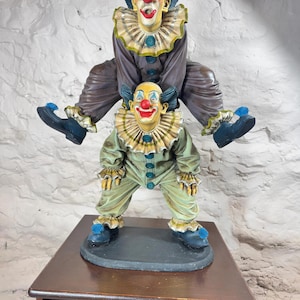 May include: A decorative figurine of two clowns, one perched on top of the other. The top clown is seated, the bottom one standing. Both have painted faces with red noses, blue hair, and ruffled collars. They wear colourful outfits. The figurine is on a wooden surface.