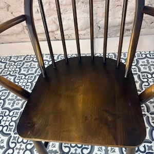May include: A dark brown wooden chair with a curved back and armrests. The chair has a solid seat and multiple vertical spindles for back support. The wood grain is visible, and the chair appears to be vintage or antique.