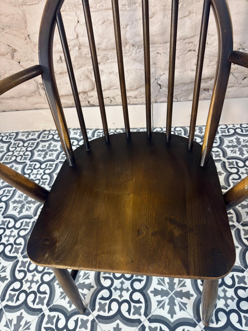 May include: A dark brown wooden chair with a curved back and armrests. The chair has a solid seat and multiple vertical spindles for back support. The wood grain is visible, and the chair appears to be vintage or antique.