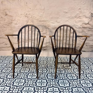 May include: Two dark wooden armchairs with a spindle back and curved top rail. The chairs have armrests and are set against a textured, light-colored wall. They sit on a patterned tile floor with a black, white, and gray design.