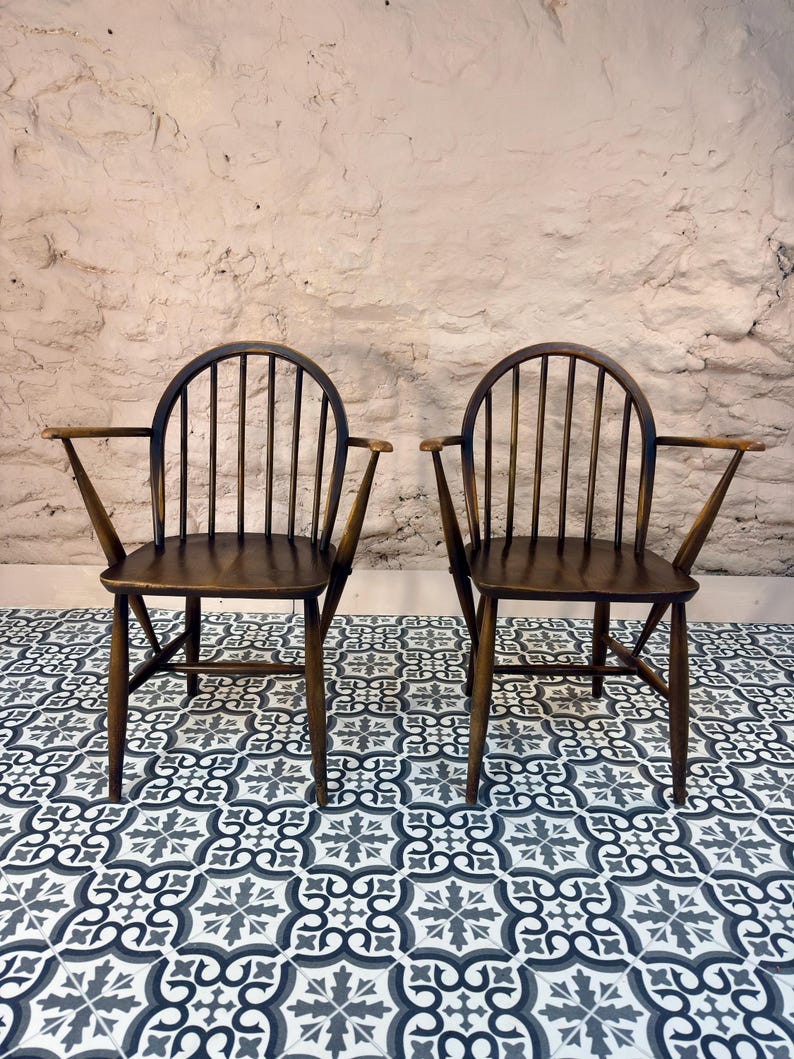 May include: Two dark wooden armchairs with a spindle back and curved top rail. The chairs have armrests and are set against a textured, light-colored wall. They sit on a patterned tile floor with a black, white, and gray design.