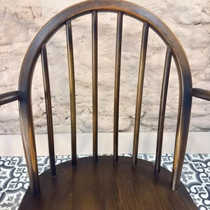May include: A dark brown wooden chair with a curved back and vertical spindles. The chair has armrests and a seat made of wood. The background is a white brick wall. The chair is a classic design.