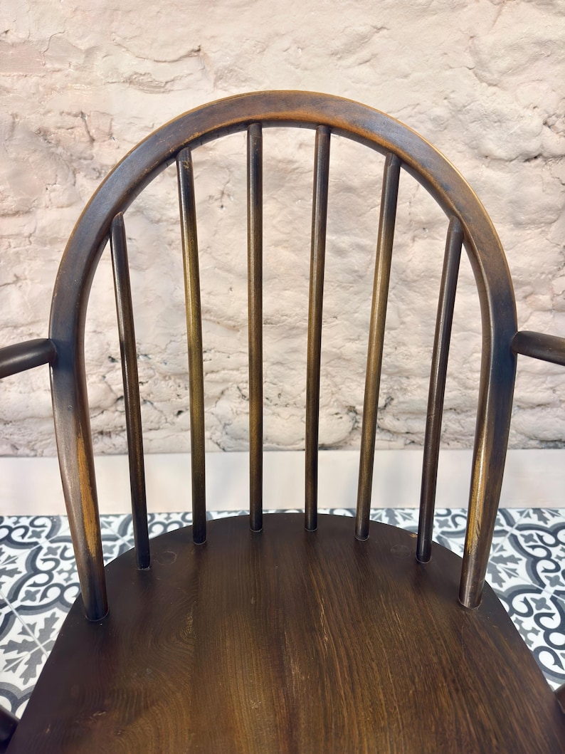 May include: A dark brown wooden chair with a curved back and vertical spindles. The chair has armrests and a seat made of wood. The background is a white brick wall. The chair is a classic design.
