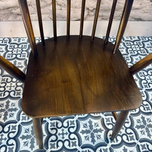 May include: A dark brown wooden chair with a curved back and armrests. The chair has a solid seat and multiple vertical spindles. The wood grain is visible, and the chair appears to be vintage or antique.