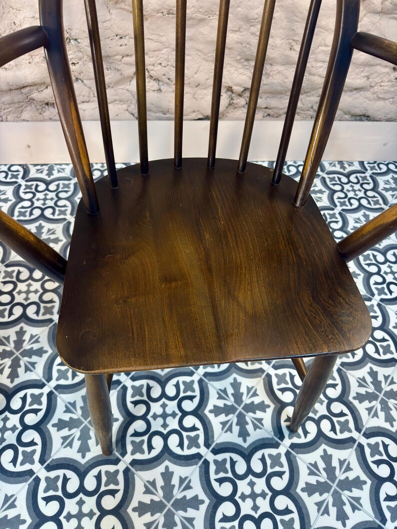May include: A dark brown wooden chair with a curved back and armrests. The chair has a solid seat and multiple vertical spindles. The wood grain is visible, and the chair appears to be vintage or antique.