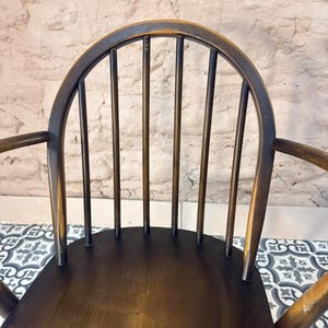 May include: A dark brown wooden chair with a curved back and armrests. The chair has a spindle back design with vertical rods. The seat is a solid, dark brown color. The chair is set against a textured, light-colored wall.