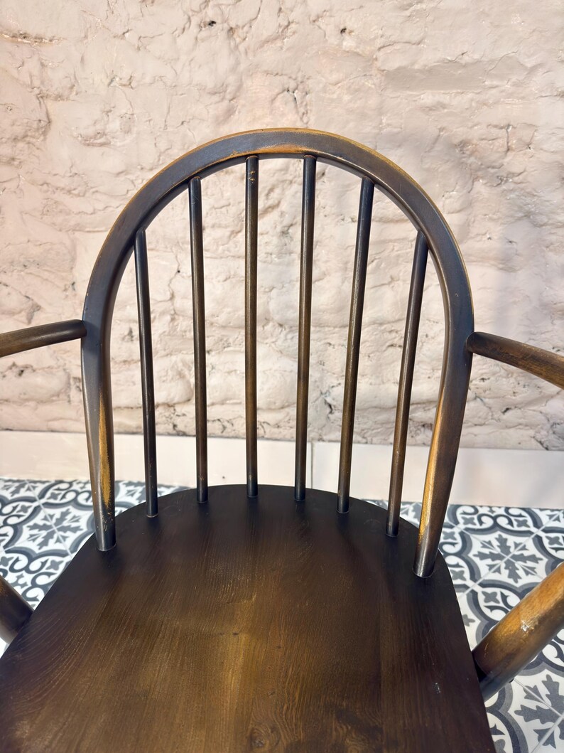 May include: A dark brown wooden chair with a curved back and armrests. The chair has a spindle back design with vertical rods. The seat is a solid, dark brown color. The chair is set against a textured, light-colored wall.