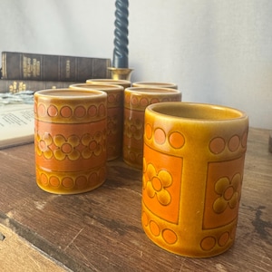May include: A collection of six vintage ceramic tumblers in orange and brown hues. The tumblers showcase a floral and geometric pattern. A lit black candle is visible in the background. The scene evokes a retro aesthetic.