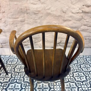 May include: A vintage wooden chair with a curved back and armrests. The chair has a dark brown seat and back supports, with a lighter brown frame. The chair is set against a white brick wall and a patterned tile floor.