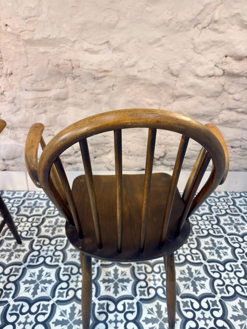 May include: A vintage wooden chair with a curved back and armrests. The chair has a dark brown seat and back supports, with a lighter brown frame. The chair is set against a white brick wall and a patterned tile floor.