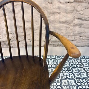 May include: Close-up of a vintage wooden chair with a curved back and armrest. The chair has a dark brown finish with visible wood grain. The back features vertical spindles. The armrest is smooth and rounded.