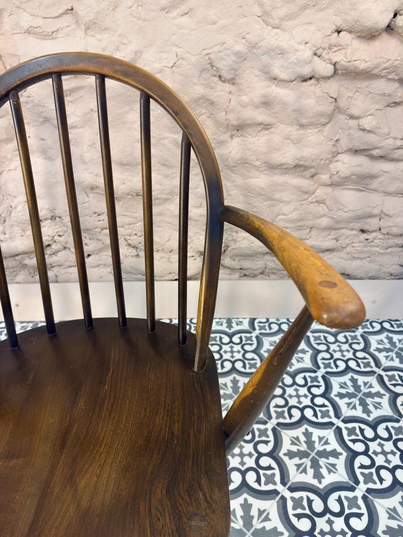 May include: Close-up of a vintage wooden chair with a curved back and armrest. The chair has a dark brown finish with visible wood grain. The back features vertical spindles. The armrest is smooth and rounded.