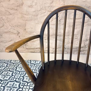May include: A vintage wooden chair with a dark brown finish. The chair features a curved back with vertical spindles and armrests. The seat is a solid piece of wood. The chair is set against a textured white wall and a patterned tile floor.