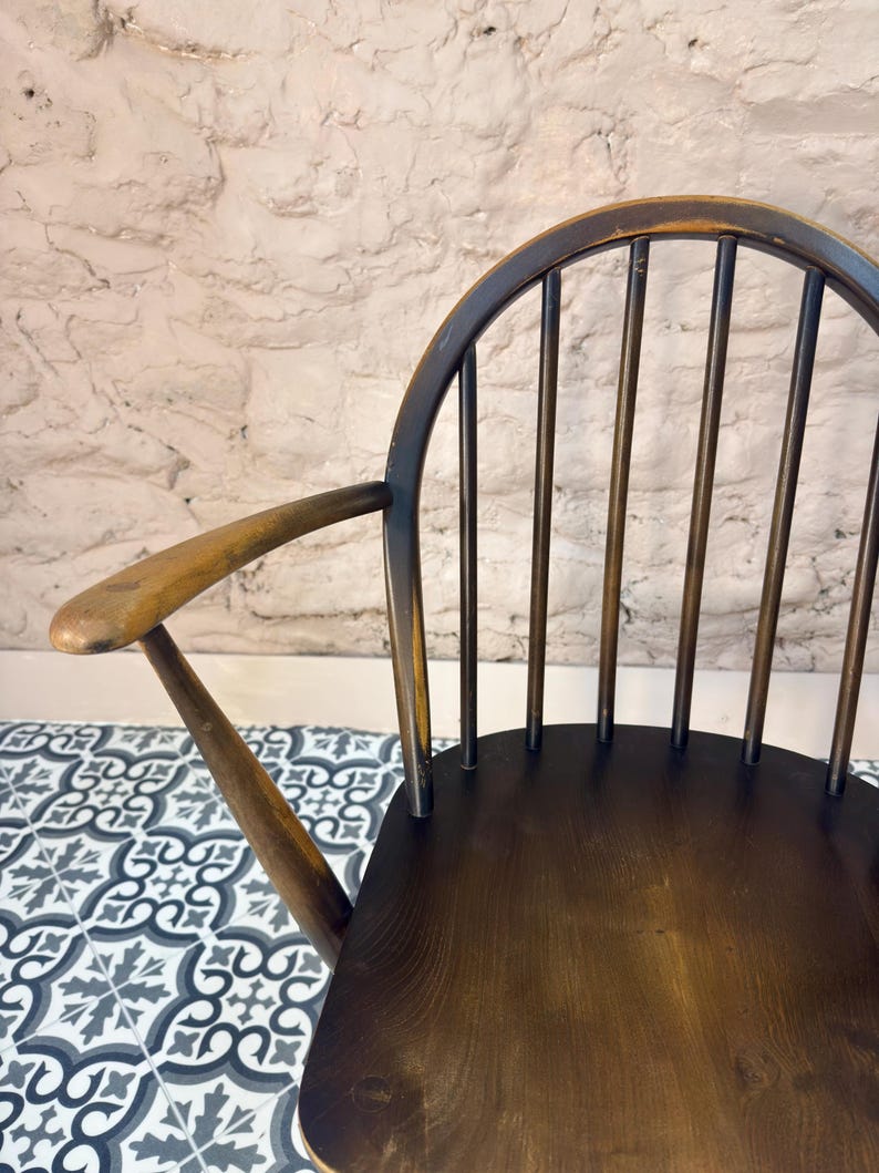 May include: A vintage wooden chair with a dark brown finish. The chair features a curved back with vertical spindles and armrests. The seat is a solid piece of wood. The chair is set against a textured white wall and a patterned tile floor.