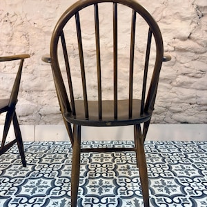 May include: A dark brown wooden chair with a curved back and vertical spindles. The chair has armrests and four legs. The chair is set against a white brick wall and sits on a patterned tile floor.