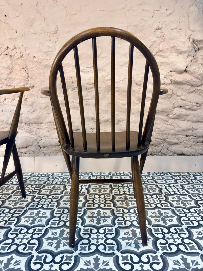 May include: A dark brown wooden chair with a curved back and vertical spindles. The chair has armrests and four legs. The chair is set against a white brick wall and sits on a patterned tile floor.