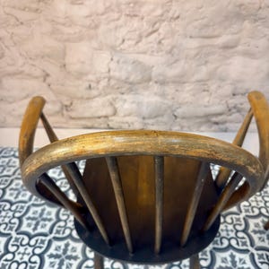 May include: Two antique wooden chairs with curved backs and spindle supports. The chairs have a distressed, aged appearance with a warm, brown finish. The seat is a dark brown color. The background is a white brick wall and a patterned tile floor.