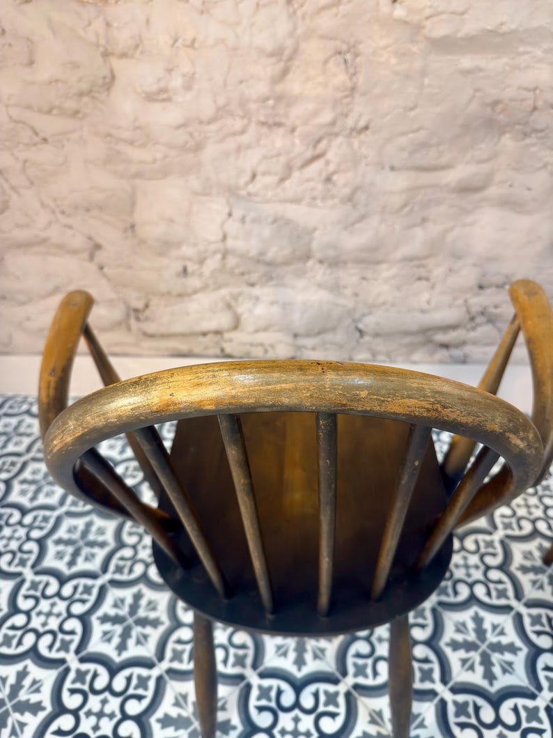 May include: Two antique wooden chairs with curved backs and spindle supports. The chairs have a distressed, aged appearance with a warm, brown finish. The seat is a dark brown color. The background is a white brick wall and a patterned tile floor.