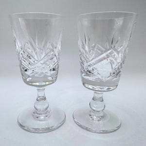 May include: A pair of clear crystal wine glasses, showcasing detailed cut glass designs. Each glass has a circular base, a stem with a rounded feature, and a flared bowl. These glasses are suitable for serving wine.