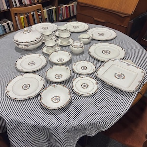 May include: A collection of vintage white porcelain dishware with a floral and gold rim design. The set includes plates, bowls, cups, saucers, a sugar bowl, a serving dish, and a covered serving bowl. The dishes are arranged on a striped tablecloth.
