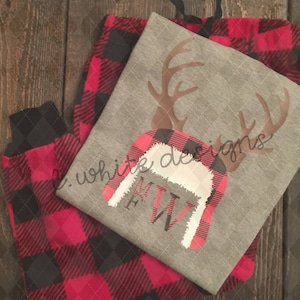 May include: Red and black plaid pajama pants with a gray long-sleeved shirt featuring a reindeer with antlers and a red and white plaid hat with the letters "FW" on it. The shirt also has the text "e. white designs" on it.