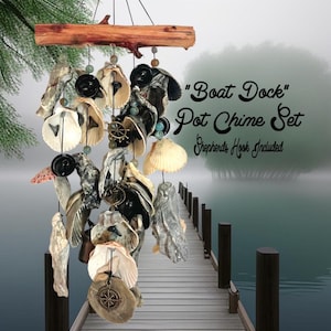 May include: A wind chime featuring a horizontal piece of driftwood at the top, from which hang multiple strings of oyster shells, black circular beads, and small metal nautical charms. The text "Boat Dock" Pot Chime Set Shepherds Hook Included is visible.