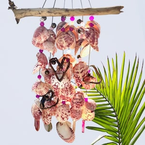 May include: A seashell wind chime with pink and white shells, strung with purple beads, hangs from a piece of driftwood. The shells are arranged in multiple strands, creating a decorative mobile. A green palm frond is in the background.