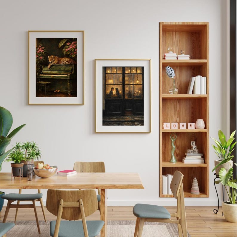 May include: Two framed art prints hang on a white wall. One features a leopard on a piano, the other depicts a couple in a doorway. A wooden bookshelf displays books and decorative items. A wooden dining table and chairs are in the foreground.