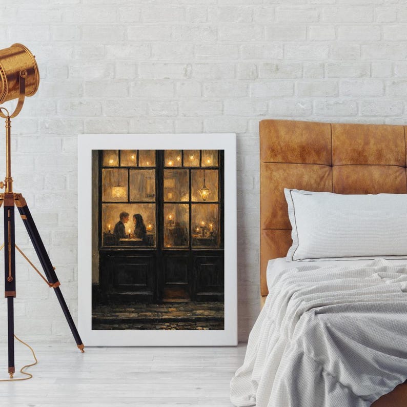 May include: A framed art print depicts a romantic scene of a couple dining in a warmly lit restaurant. The artwork, in a white frame, is leaning against a white brick wall. A brown leather headboard and a bed with white linens are in the background.