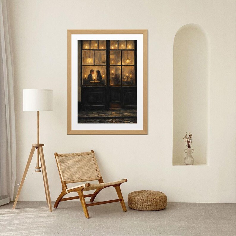 May include: A framed print depicts a cozy scene of a couple in a warmly lit cafe. The artwork, in a wooden frame, features a dark door and windows with soft, golden light, creating a romantic ambiance. The print is displayed in a neutral-toned room.